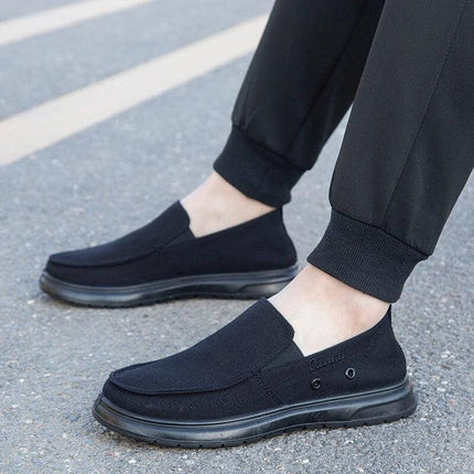 Zapatos de lona casuales negros para hombre, ideales para caminar, mostrando diseño moderno con pantalones oscuros sobre pavimento.