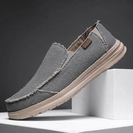 Zapato casual de hombre de lona gris transpirable exhibido en un pedestal blanco.